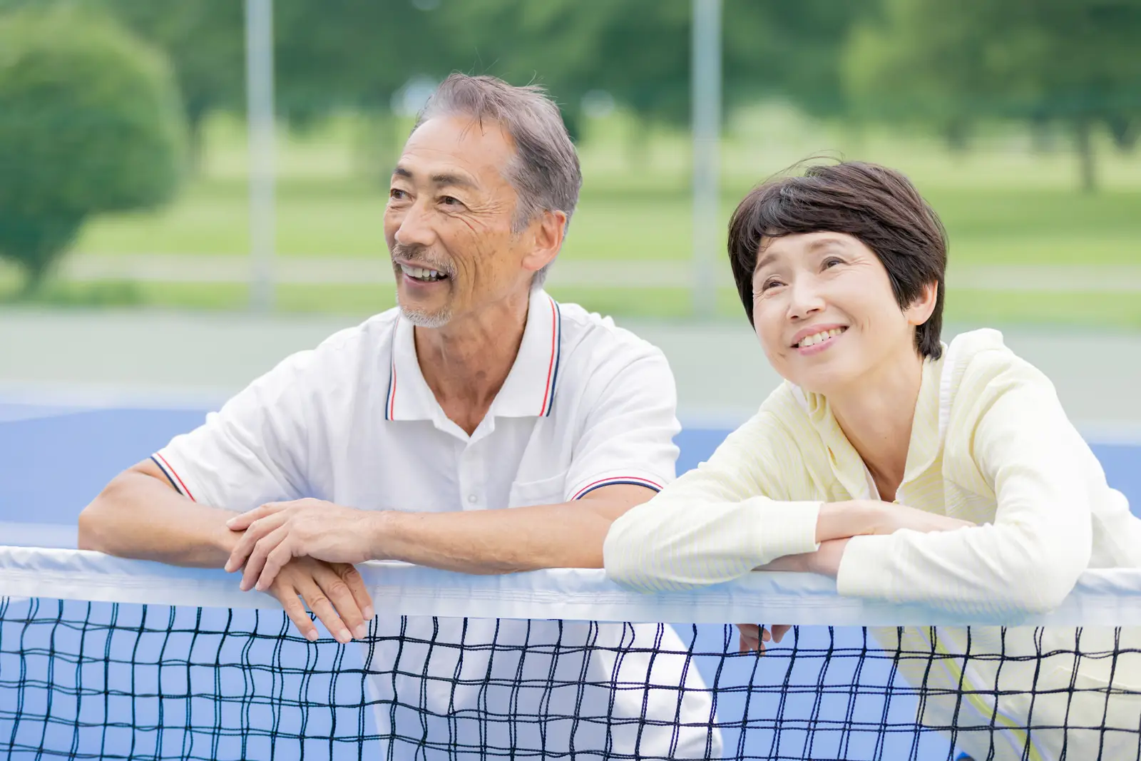 テニスコートのネットに寄りかかり、笑顔で遠くを見る50〜60代の夫婦（イメージ）